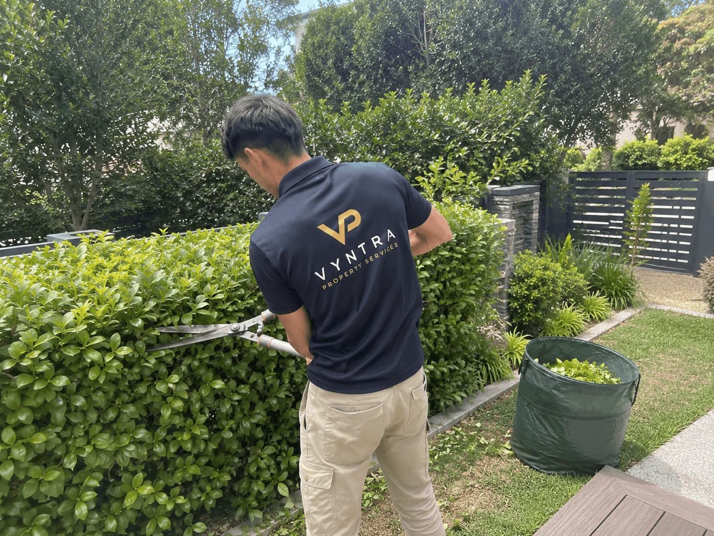 A Vyntra gardener trimming a hedge with shears, green waste bag alongside